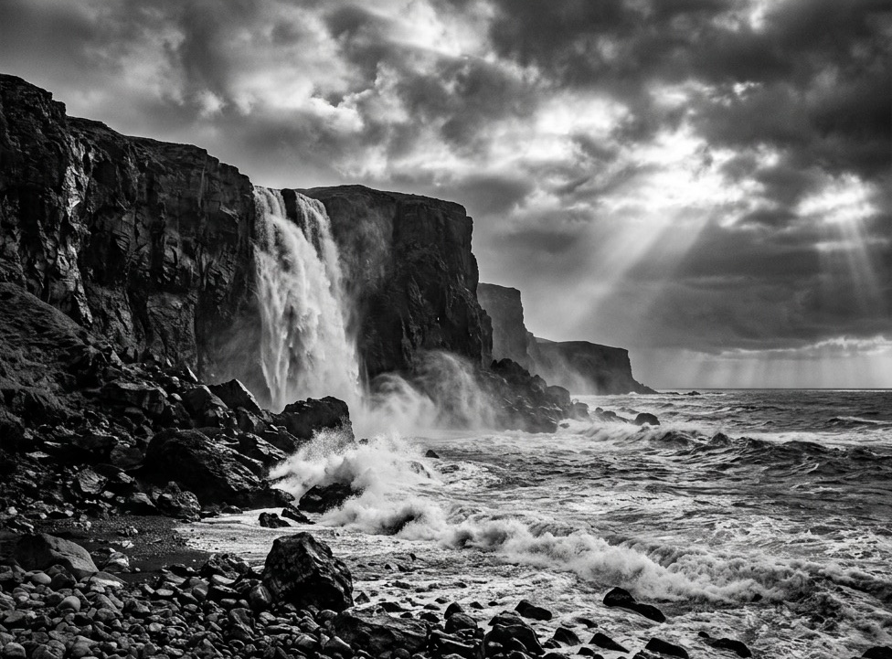 Black-and-white fine-art photograph presented on a gallery matte: a waterfall pours off basalt sea cliffs into storm surf while crepuscular rays break through clouds